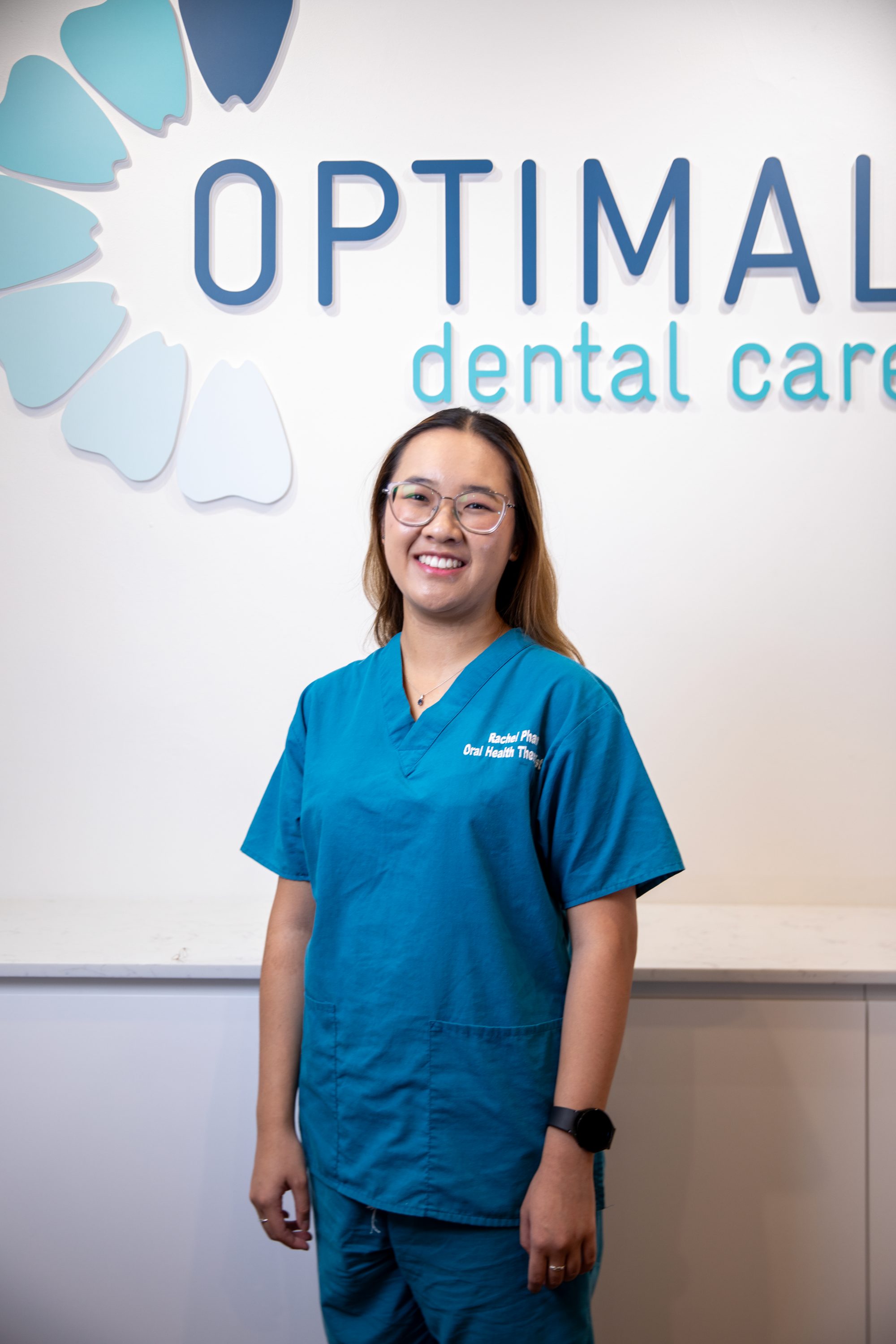 Rachel Pham, Oral Health Therapist at Optimal Dental Care, in teal scrubs by the practice wall logo