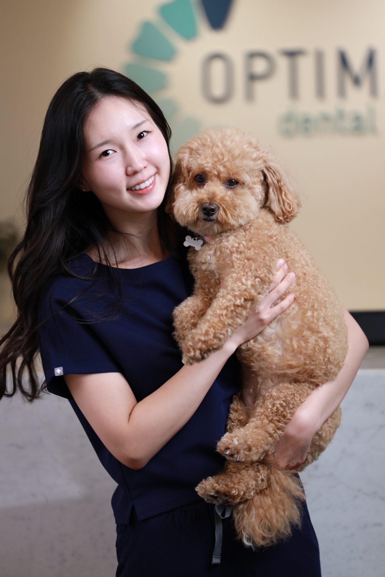 A member of the Optimal Dental Care team in navy scrubs smiling while holding her small dog by the wall logo
