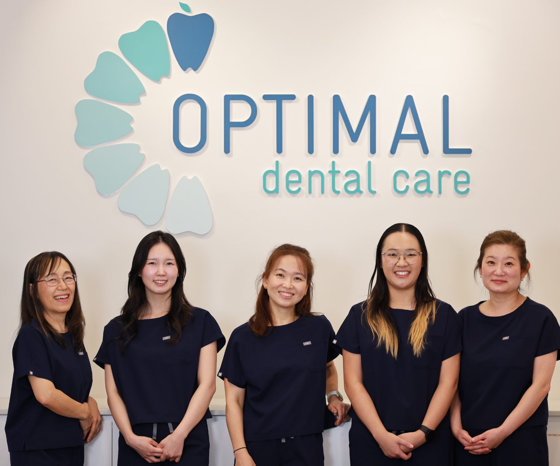 The Optimal Dental Care team in navy scrubs in front of the practice wall logo at the Woollahra clinic